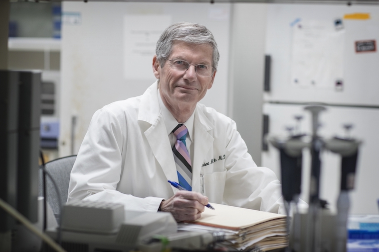 Robert Carey wears a white coat and holds a pen as he smiles at the camera.