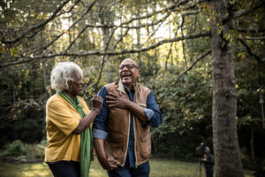 A senior man and woman laugh outside amid green trees.