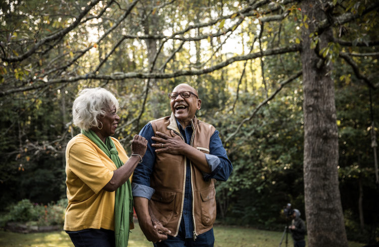 A senior man and woman laugh outside amid green trees.