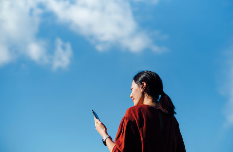 A woman looks at her cell phone in front of a blue sky.
